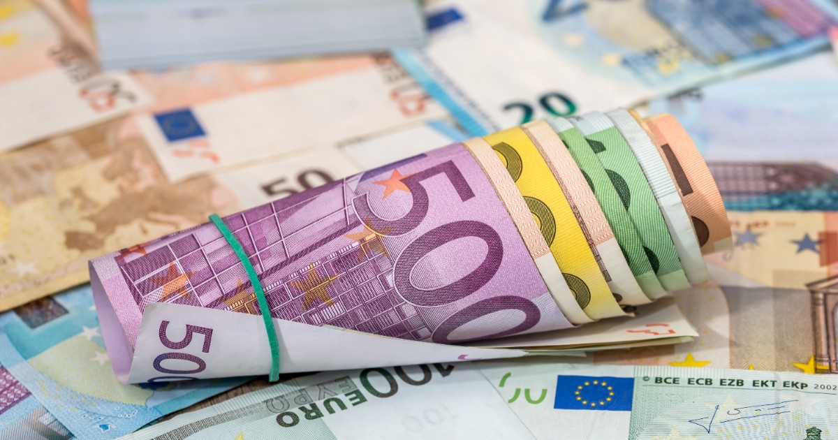 Imaginea prezintă un sul de bancnote euro cu valori mari legat cu un elastic de cauciuc, așezat pe un fundal format din alte bancnote împrăștiate.