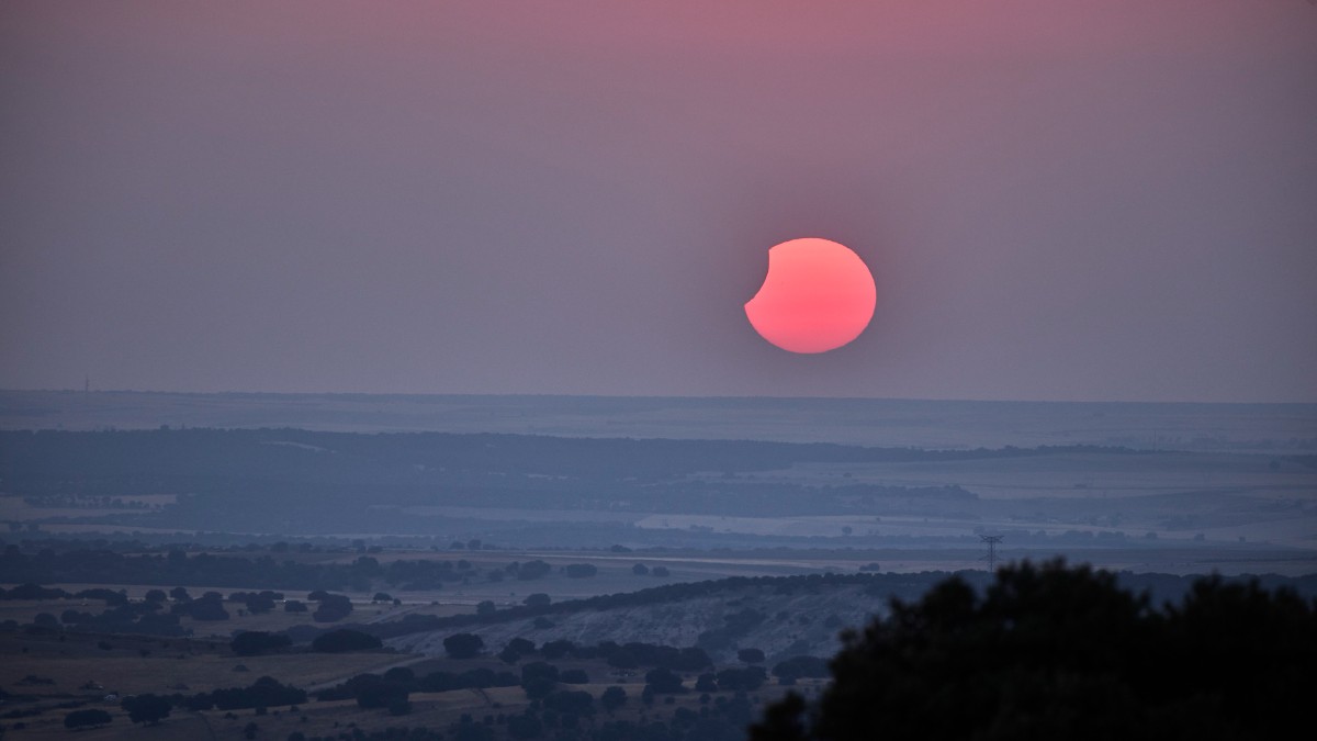 În imagine se vede o eclipsă solară parțială, cu discul Soarelui acoperit în parte de Lună și colorat în nuanțe roșiatice. În prim-plan se întinde un peisaj rural vast, surprins în lumina slabă și atmosferică a fenomenului astronomic.