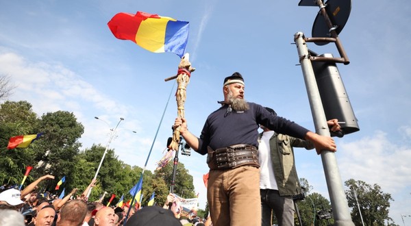 Un bărbat cu barbă și brâu din piele, purtând un toiag decorat și un steag al României, urcat la înălțime deasupra unei mulțimi de protestatari.