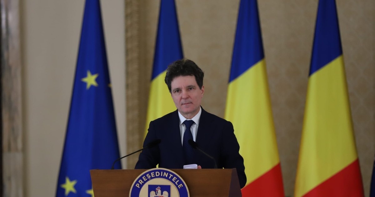Președintele Nicușor Dan susține o declarație oficială de la pupitrul Administrației Prezidențiale, având în fundal drapelul României și steagul Uniunii Europene. Președintele Nicușor Dan adoptă o ținută formală, specifică protocolului de stat, purtând un costum de culoare închisă (bleumarin sau negru), o cămașă albă și o cravată asortată, într-o nuanță închisă de albastru.