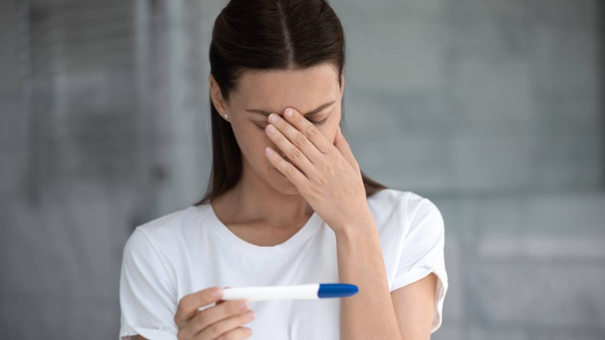 O femeie cu un test de sarcină în mână, și-a dus o mână la cap și privește tristă spre rezultatul indicat