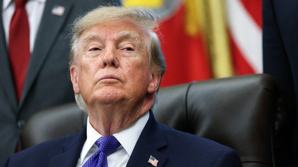 Donald Trump privind în sus, îmbrăcat într-un costum închis la culoare, cămașă albă, cravată albastră cu model și un pin cu drapelul Statelor Unite la rever.