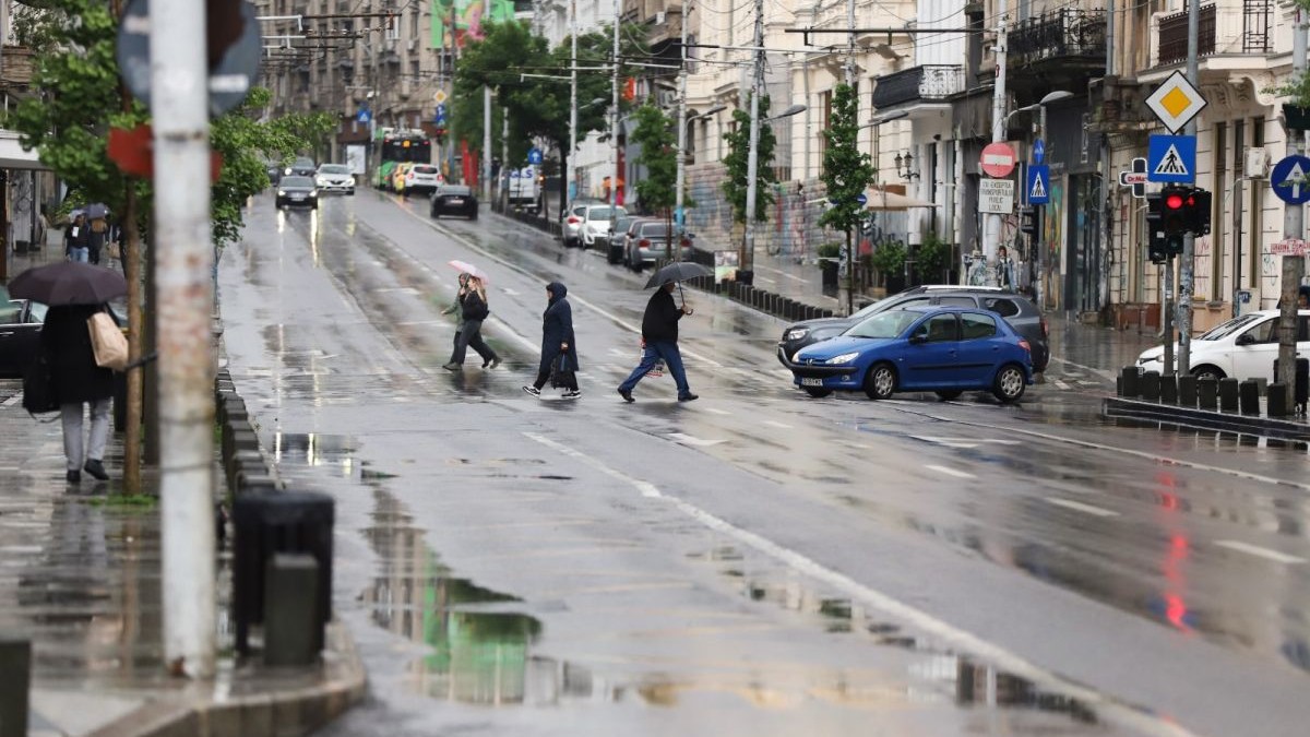 Mai mulți oameni cu umbrele în mâini traversează strada pe la trecerea de pietoni, câteva mașini sunt oprite, iar pe trotuar sunt alți oameni cu umbrele, într-o zi ploioasă de vară, zona Cișmigiu, București, 26 mai 2025