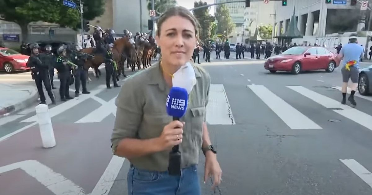 femeie blonda, in blug si camasa, cu un microfon in mana, in mijlocul unor proteste, forte de ordine in spatele ei,
