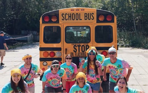 9 americani imbracati in tricouri colorate pozeaza langa un autobuz galben scolar din SUA