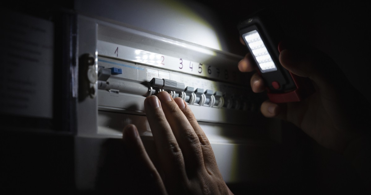Un bărbat verifică cu lumina de la telefonul mobil un panou electric într-un mediu întunecat și iluminează siguranțele și întrerupătoarele din panou.