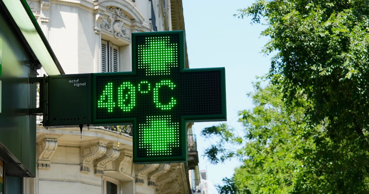 Plan apropiat cu o firmă luminoasă verde în formă de cruce a unei farmacii, care afișează o temperatură caniculară de 40 de grade Celsius, pe fundalul unei clădiri cu arhitectură clasică și al unor copaci verzi.