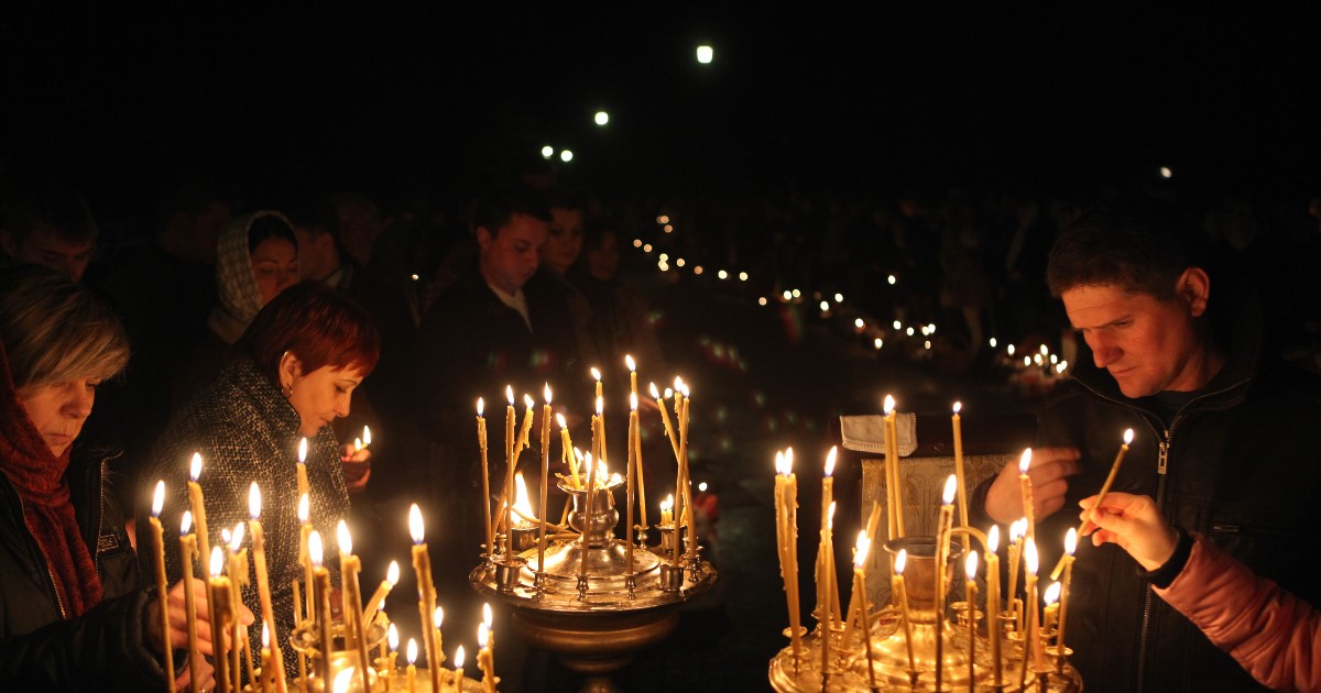 Mai multe persoane aprind lumânări într-un cadru religios, în timpul nopții, creând o atmosferă solemnă și liniștită. Flăcările lumânărilor luminează fețele credincioșilor, sugerând un moment de reculegere și rugăciune.