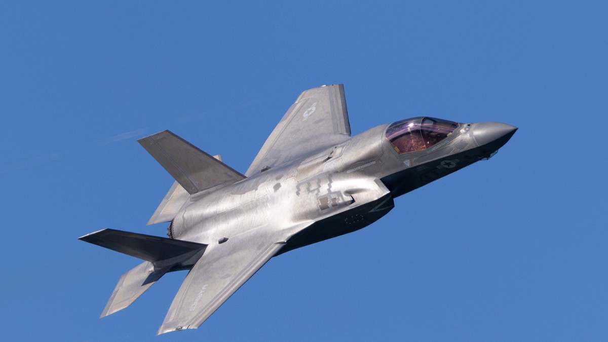 Un avion militar modern, de tipul F-35, zboară pe un cer senin, de un albastru intens. Aeronava este de culoare gri metalic și are o formă specifică de avion de vânătoare stealth, fiind surprinsă în plin zbor.