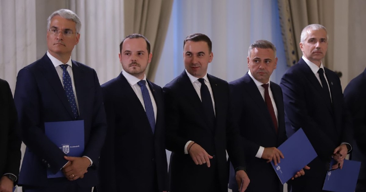 Miniștrii din Guvernul Bolojan, îmbrăcați în costume negre, țin o mapă albastră în mână, sunt aliniați pentru o poză.