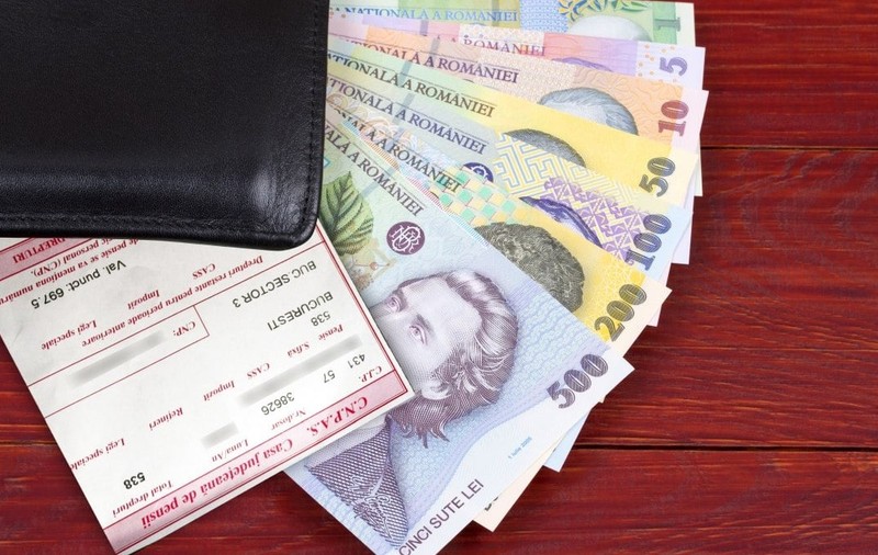 Mai multe bancnote românești ies dintr-un portofel al cărui colț negru se vede pe o masă, iar deasupra banilor este un cupon de pensie