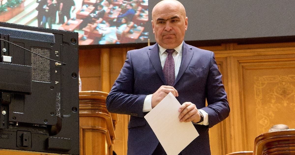 Imaginea îl înfățișează pe Ilie Bolojan purtând un costum bleumarin și cravată, ținând niște documente în mână, în Parlamentul României.