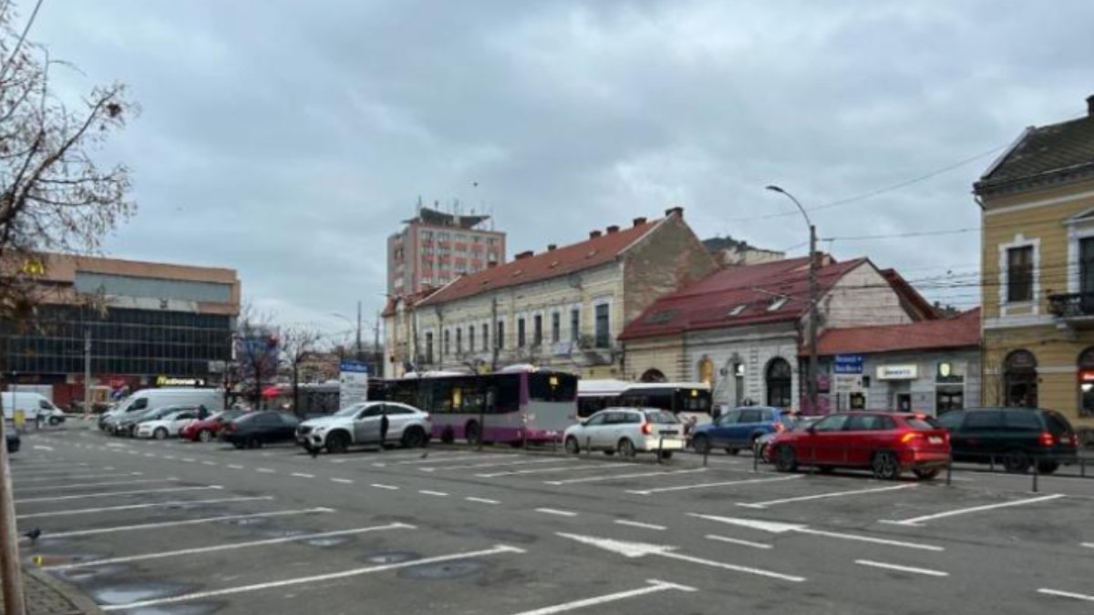 Imaginea surprinde parcarea din Piața Mihai Viteazul din Cluj-Napoca, într-o zi mohorâtă. Se pot observa mai multe mașini parcate, autobuze în stație.