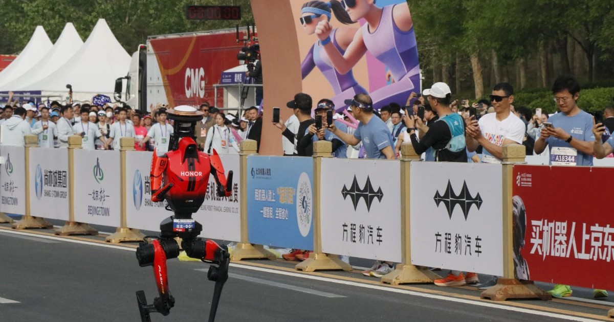 Un robot umanoid aleargă pe traseul unui semimaraton, în timp ce este urmărit de spectatori care fotografiază momentul. Evenimentul are loc într-un cadru organizat, cu bariere și bannere, sugerând o cursă oficială desfășurată în Beijing.