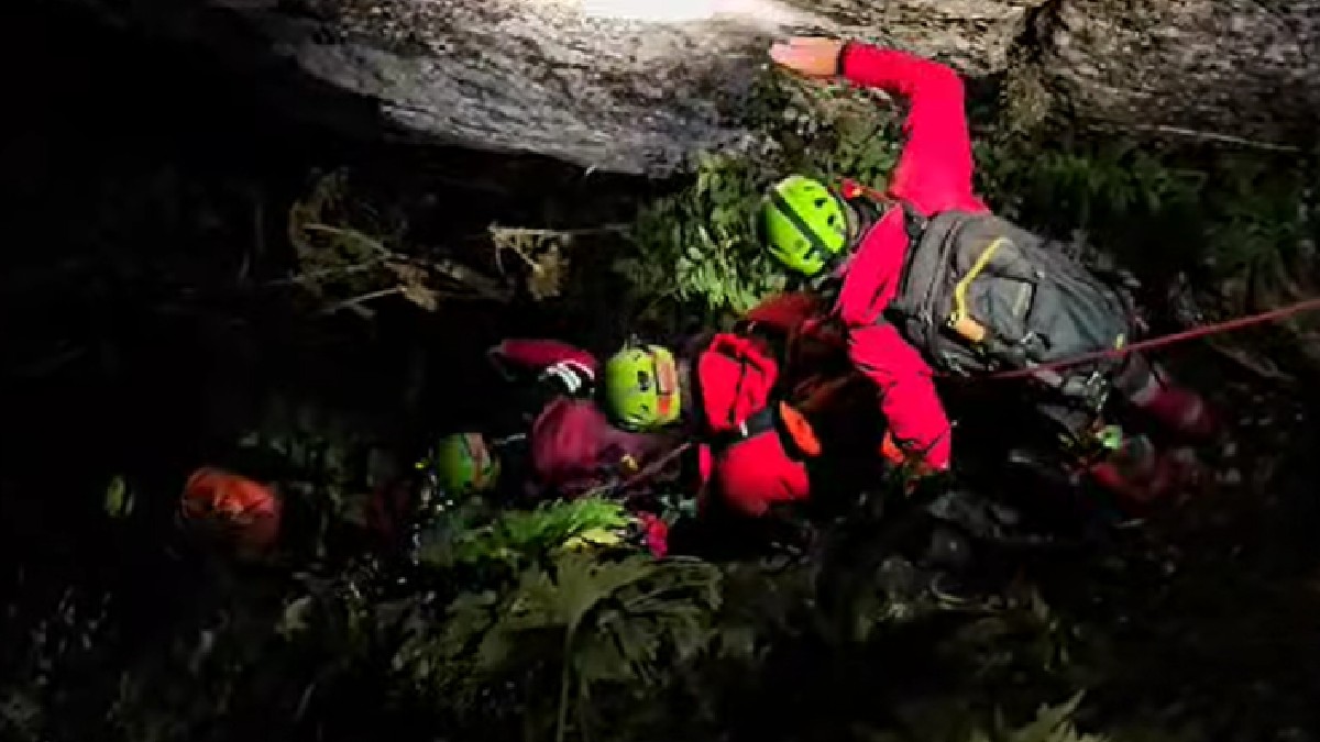Imaginea arată un grup de salvatori, salvamontiști, într-un mediu întunecat și dificil. Persoana din prim-plan poartă o geacă roșie, o cască verde deschis și un rucsac, și pare să fie implicată într-o acțiune de alpinism sau de salvare, fiind prinsă de o coardă. Lumina pare să provină de la o lanternă sau o sursă de lumină puternică, care iluminează dramatic zona. În jur se văd stânci, vegetație și alte persoane, echipate similar, ceea ce sugerează o operațiune de salvare într-un peisaj montan.