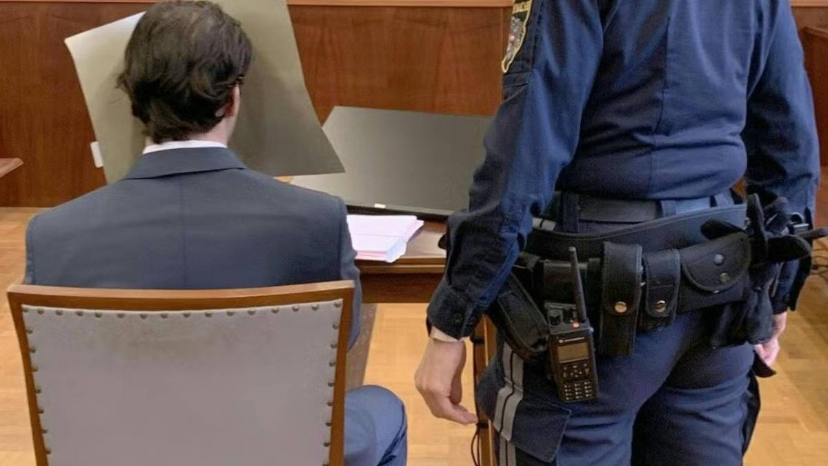 Imaginea arată un bărbat în costum, văzut din spate, stând la masa dintr-o sală de judecată. Își acoperă fața cu un paravan gri. Lângă el stă un polițist în uniformă albastră, cu echipament pe centură.
