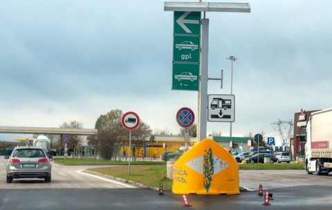 O zonă de servicii și stație de alimentare de pe o autostradă din Italia, cu panouri verzi de direcționare, indicatoare rutiere, un autoturism Volkswagen argintiu în mers și un camion staționat în dreapta.