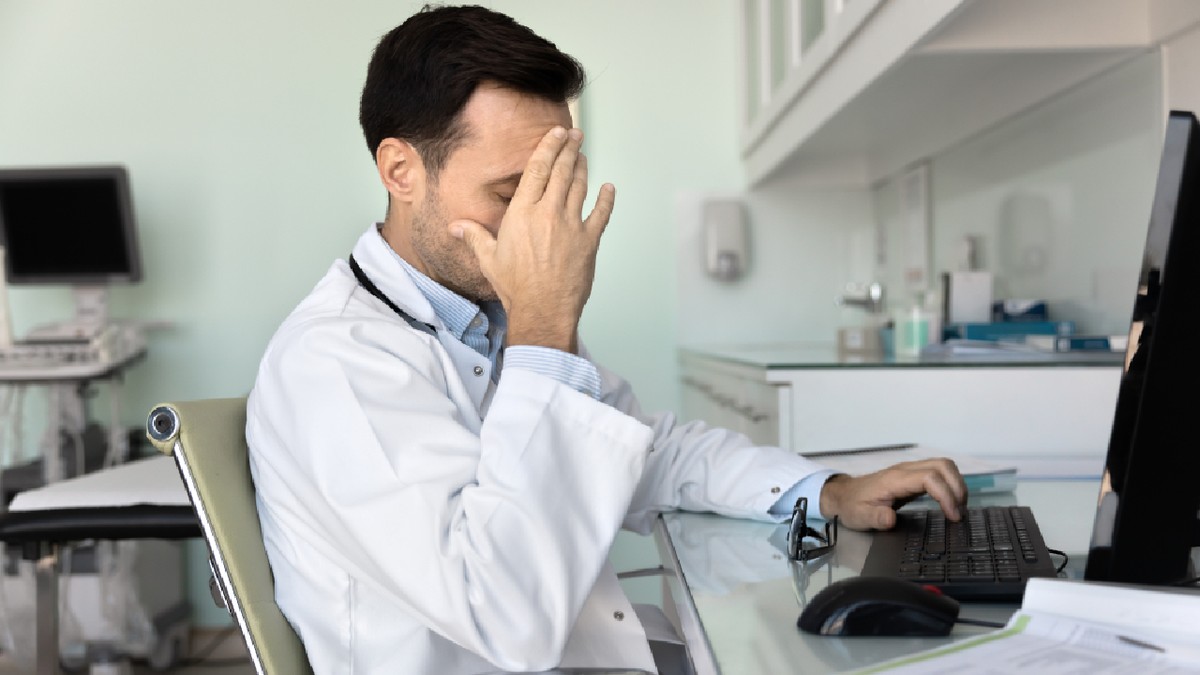 Doctorul stă la birou cu calculatorul și hârtiile, acoperă fața cu mâna, se simt epuizat, supraîncărcat, pare stresat din cauza erorilor medicale sau a diagnosticării greșite, face greșeli, se confruntă cu un caz dificil de pacient