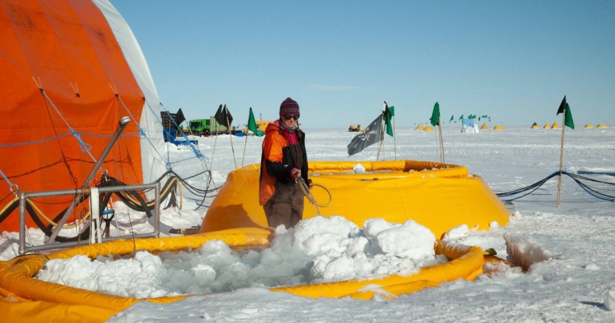 Un cercetător îmbrăcat în echipament gros de iarnă, portocaliu și negru, stă pe gheața din Antarctica și folosește un furtun care pulverizează apă fierbinte peste zăpadă. În jur se văd structuri gonflabile galbene, echipamente științifice și cabluri întinse. În fundal apar mai multe corturi colorate, dispuse pe câmpul alb de gheață, sub un cer senin.
