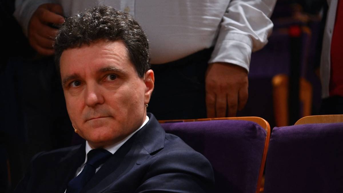 Nicușor Dan, îmbrăcat la costum, în timp ce stă pe scaun într-o sală de evenimente.