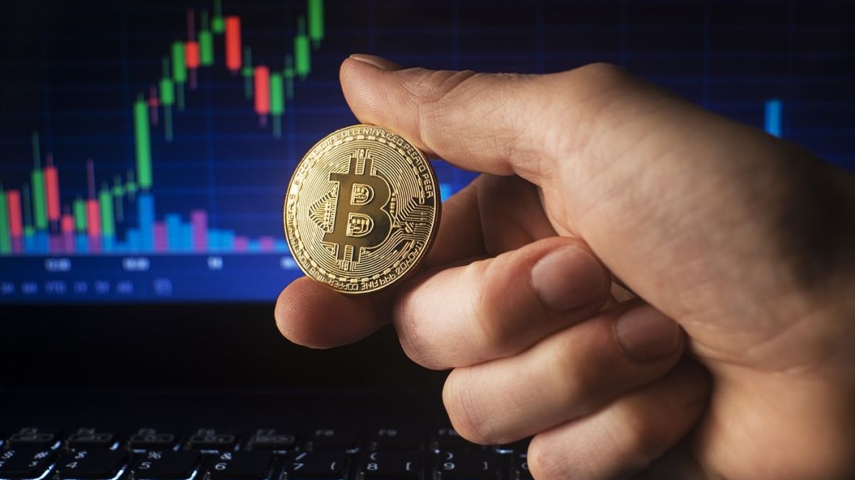 Un bărbat ține în mână o monedă bitcoin auriu pe fundalul graficului de tranzacționare pe piața bursieră