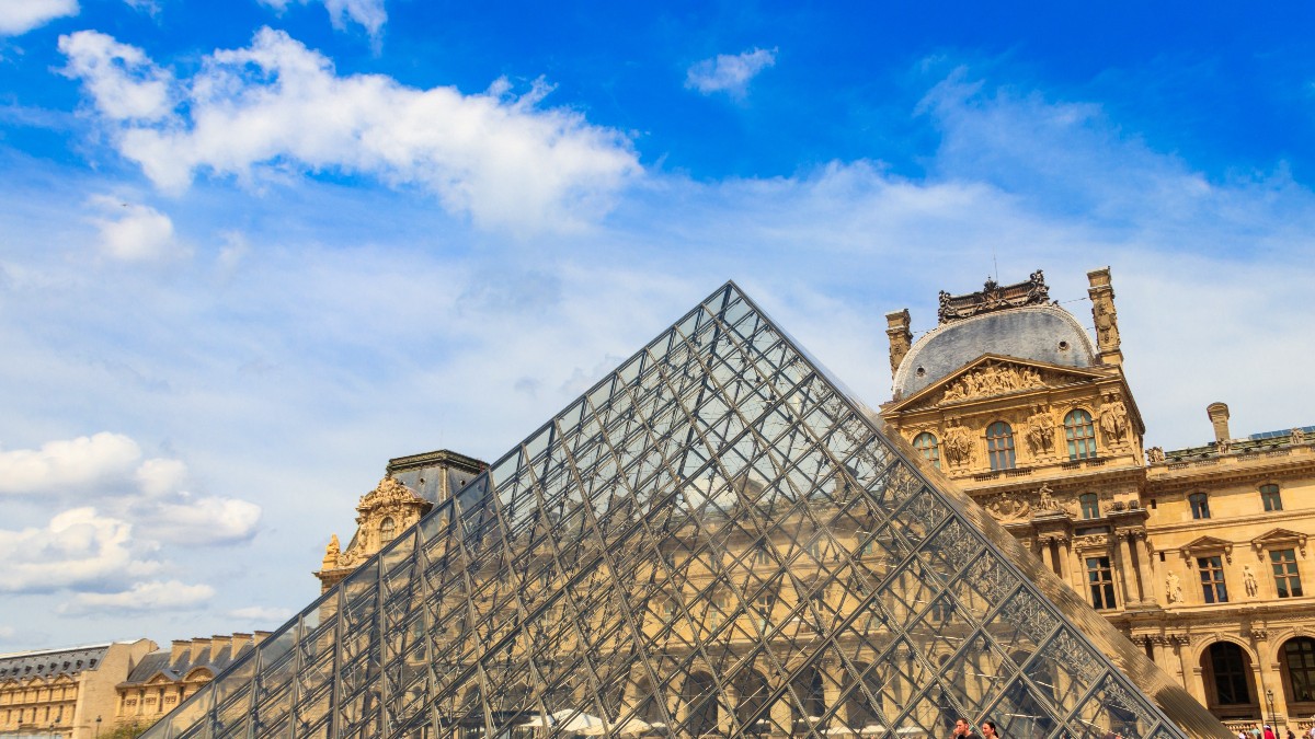 Imaginea prezintă celebra Piramidă de Sticlă din fața Muzeului Luvru din Paris, care contrastează puternic cu arhitectura clasică, masivă, a clădirilor din jur, sub un cer albastru cu nori puțini. Piramida modernă, cu structura sa metalică și panourile de sticlă, domină centrul cadrului, în timp ce la bază se pot observa câțiva vizitatori.