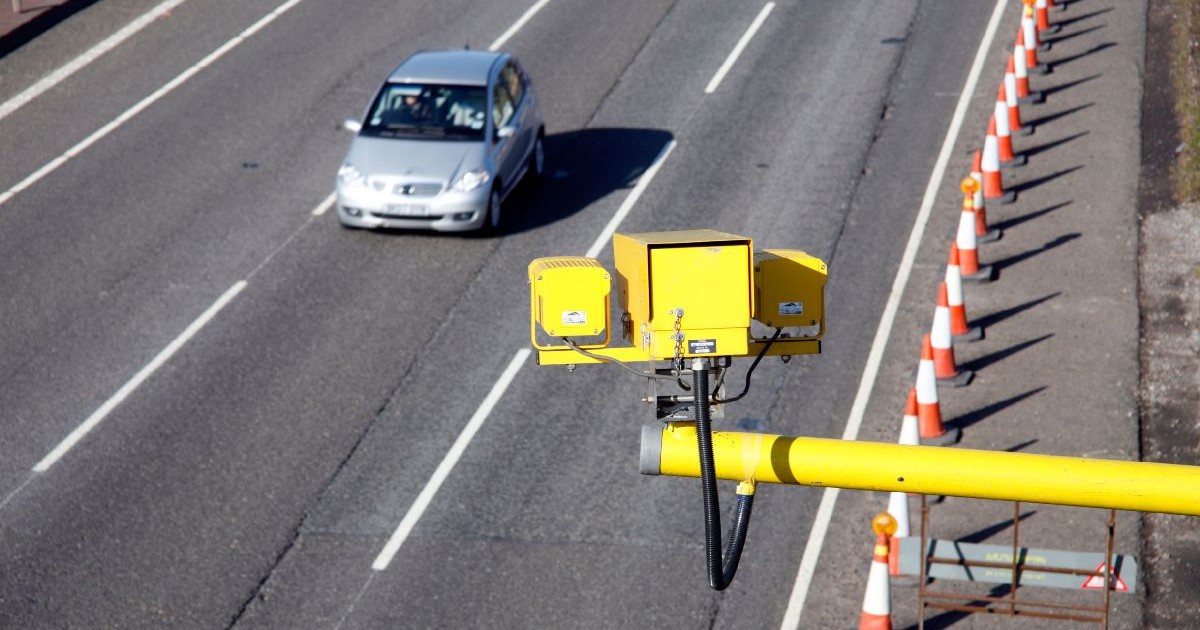 Imaginea prezintă un radar fix de culoare galbenă montat deasupra unei șosele, în timp ce în fundal se observă un autoturism gri aflat în trafic și un rând de conuri de semnalizare rutieră.