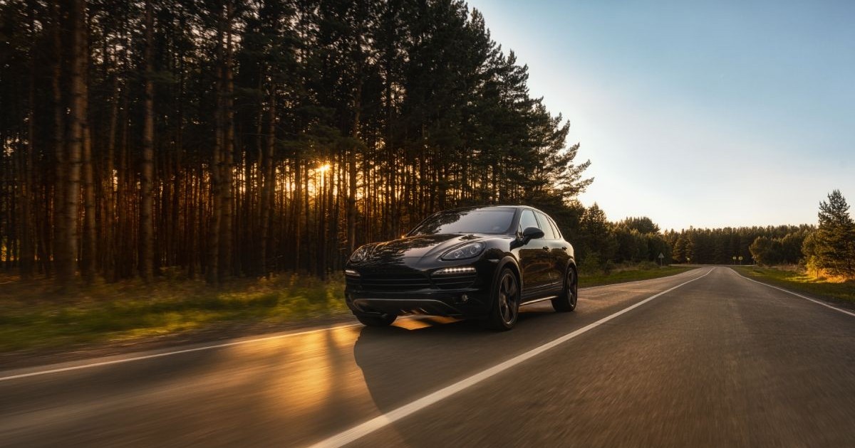 Un Porsche Cayenne se deplasează pe un drum pe lângă pădure, soarele se zărește printre copaci
