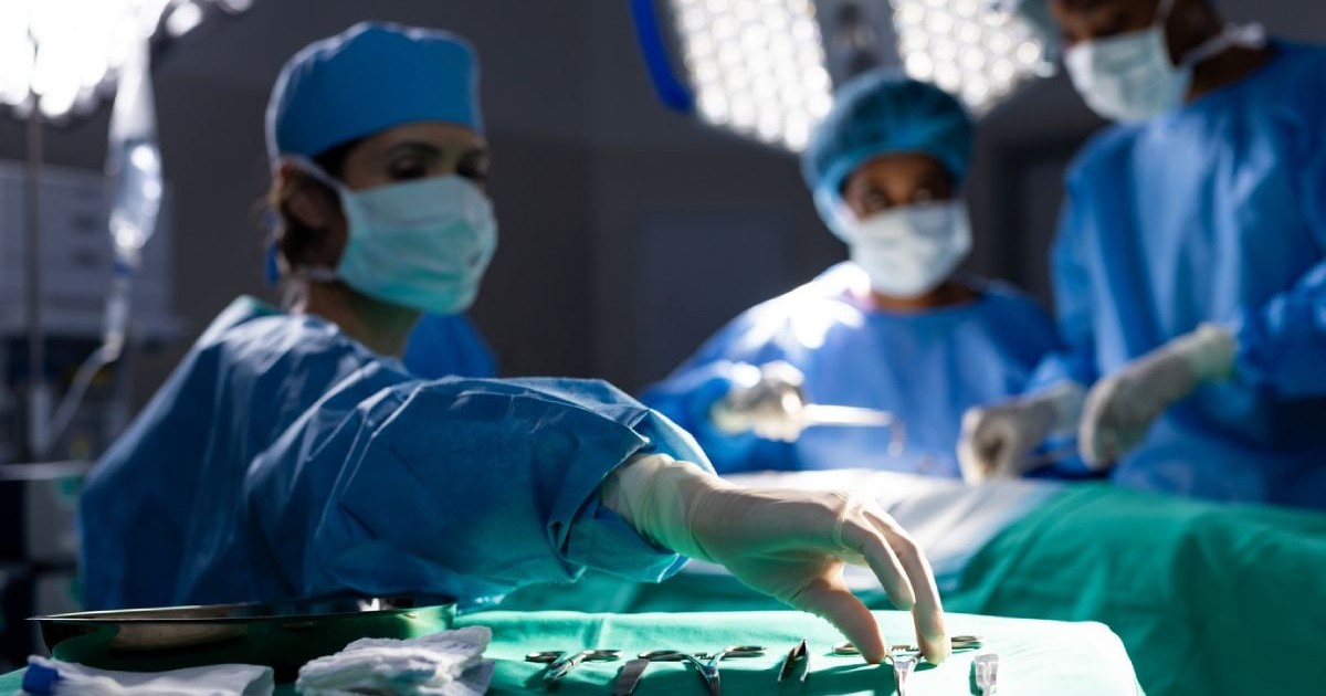 Trei medici în costume sterile albastre, cu masti pe față in timpul unei operații, in afat se vede instrumentarul steril