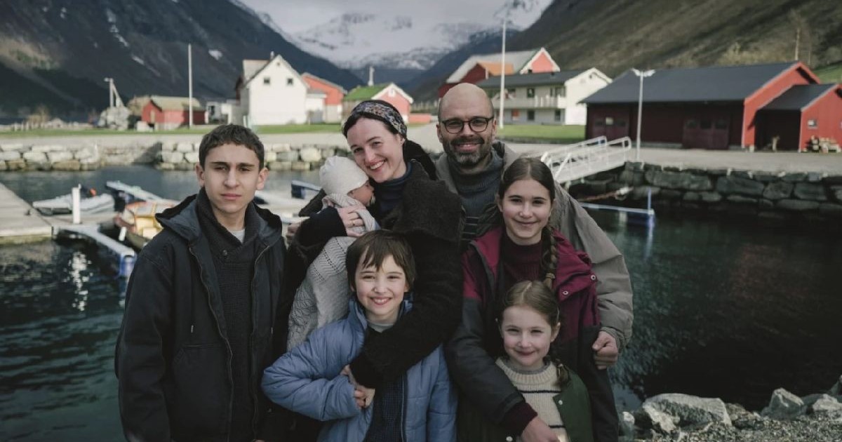 Scenă din filmul „Fjord” al lui Cristian Mungiu: un grup de persoane, adulți și copii, zâmbind în timp ce pozează împreună în apropierea unui mal de apă, având în fundal case colorate și munți.
