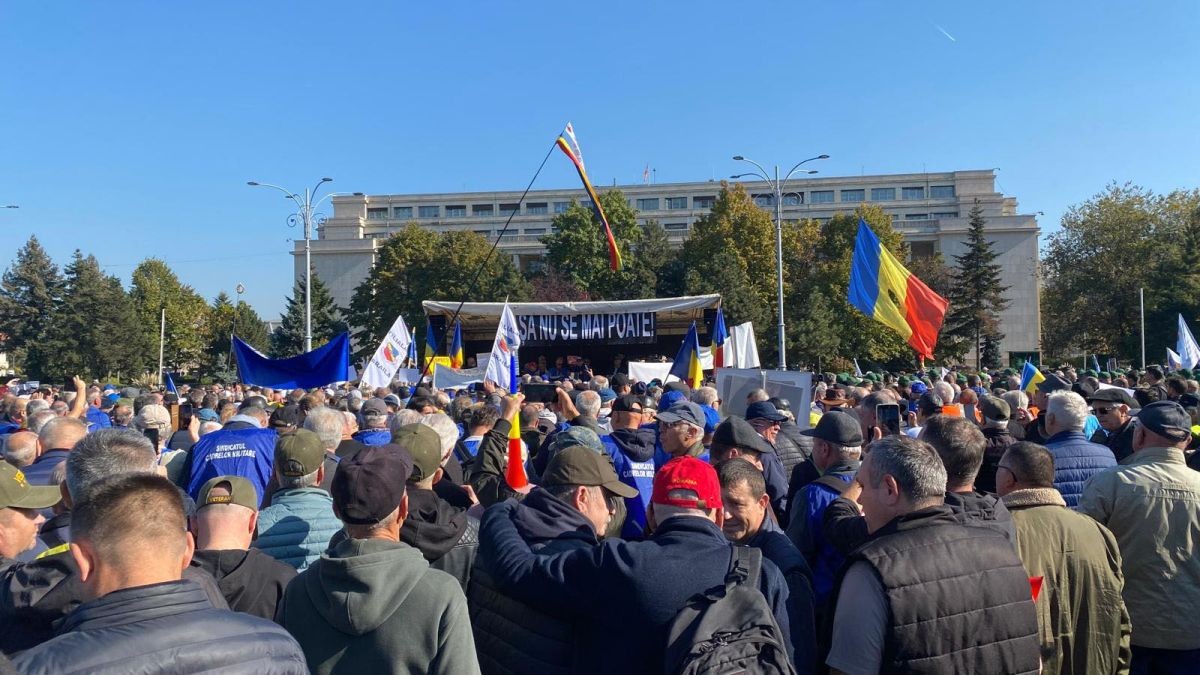 Sute de oameni purtând bannere și steaguri participă la un protest în Piața Victoriei din București, martie 2025