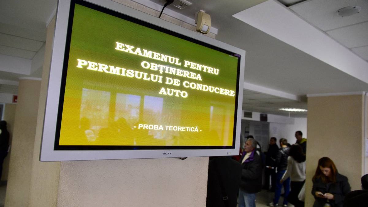 monitor pe care este afisat mesajul examen auto permis intr-o sala de asteptare unde sunt mai multi oameni