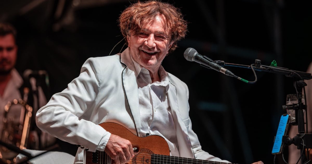 Un muzician cântă la chitară pe scenă, zâmbind în timp ce se află în fața unui microfon. Este îmbrăcat într-un costum alb, iar în fundal se observă alți membri ai formației și instrumente muzicale.