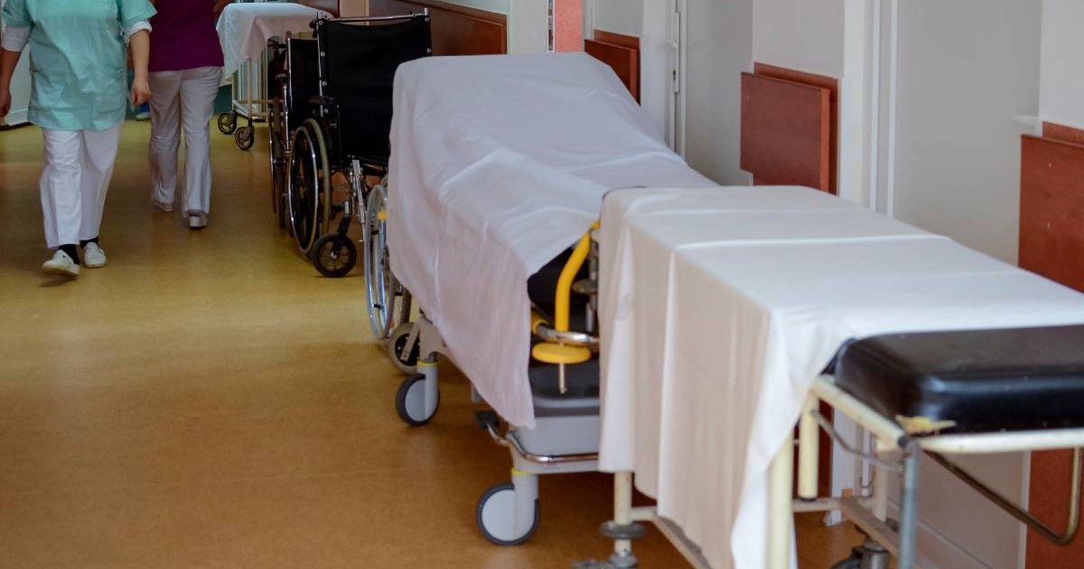 Hol de spital, cu doua paturi in dreapta acoperite cu cearșafuri albe, in stanga se vad picioarele unor cadre medicale