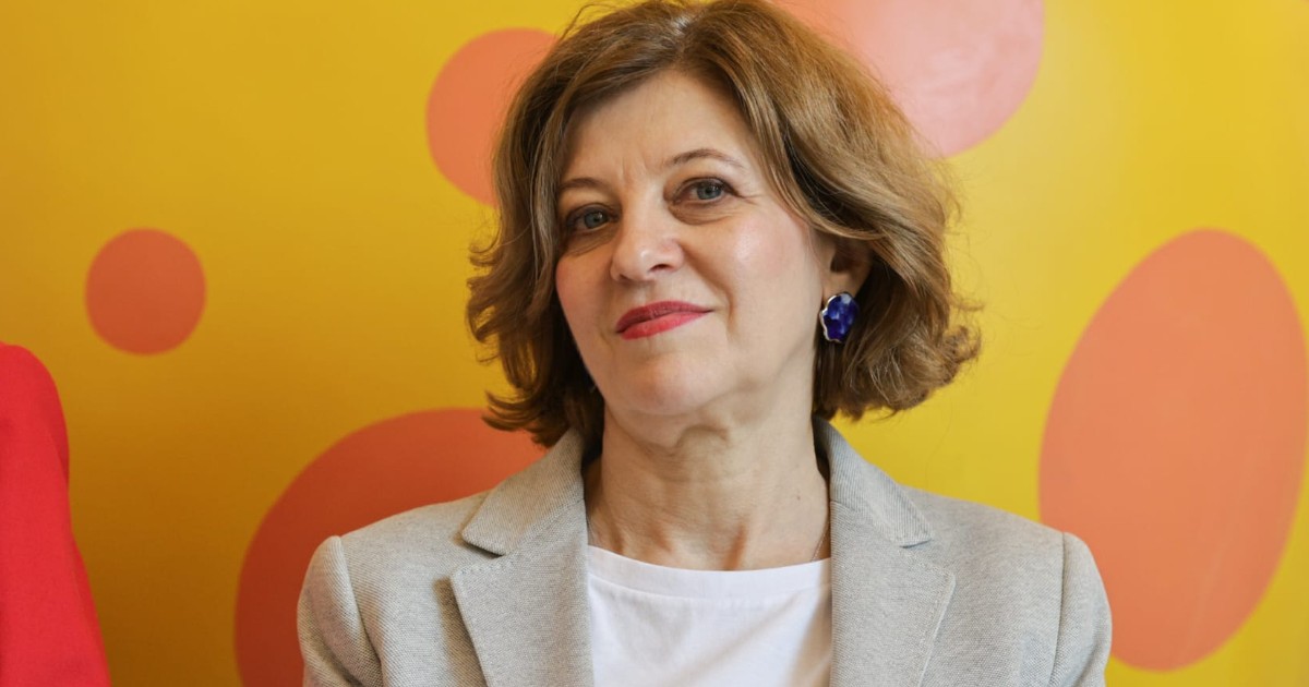 Oana Gheorghiu îmbrăcată în sacou bej și tricou alb. În spatele ei este un perete galben cu cercuri portocalii
