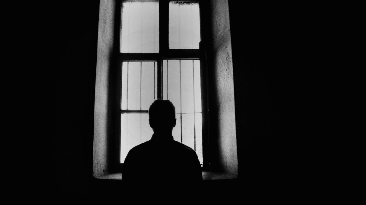Silueta unui barbat care se uita pe geam, într-o cameră întunecată.
