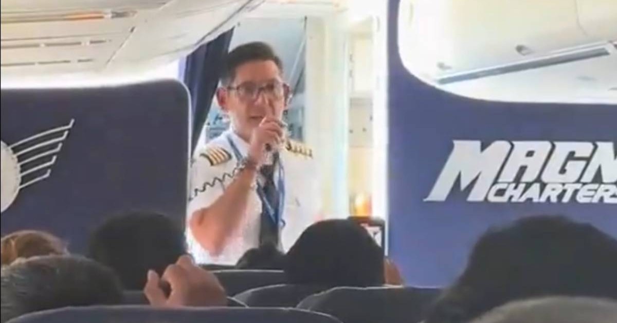 Un pilot in camasa alba si ochelari, vorbeste la sistemul de comunicare al avionului in timp ce sta in usa cabinei, se vad scaunele din avion cu capetele persoanelor din acion