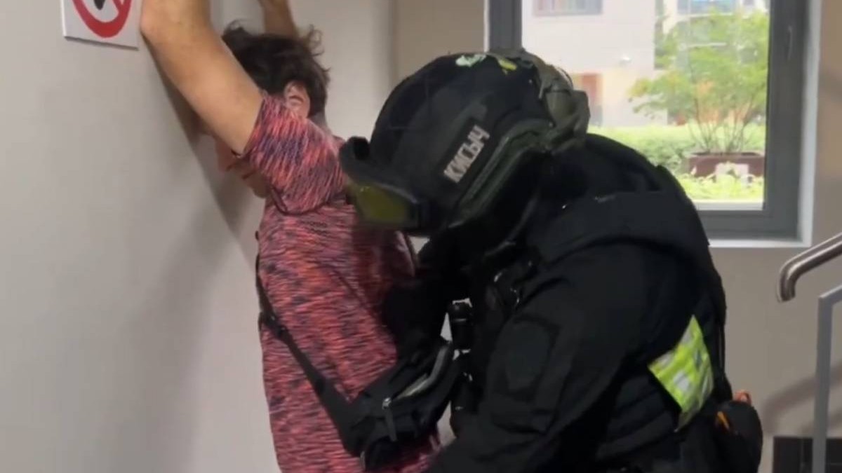 Un bărbat îmbrăcat cu un tricou roșu este percheziționat de un polițist echipat în uniformă tactică neagră, cu cască și vestă de protecție. Bărbatul stă cu fața spre perete și cu brațele ridicate, într-un hol luminos al unei clădiri.