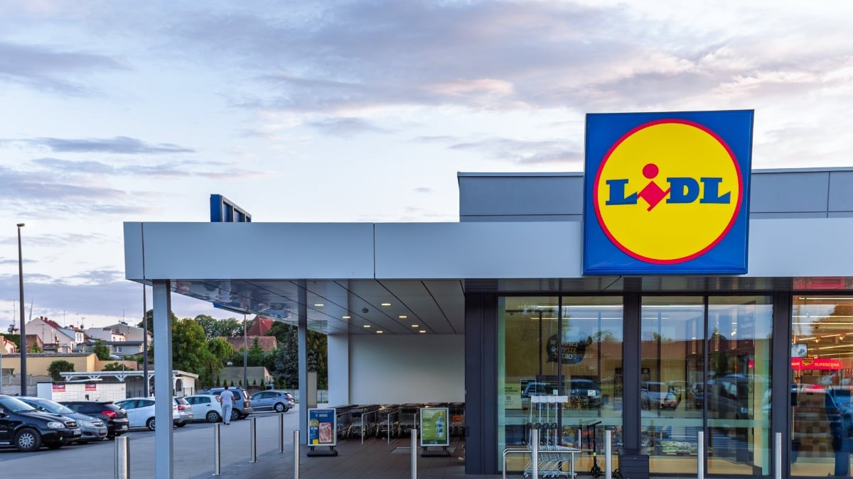 Un magazin Lidl modern este surprins din exterior, cu intrarea largă și logo-ul vizibil deasupra ușilor automate. În fața clădirii se află parcarea și zona pentru cărucioare, sugerând acces facil pentru clienți.