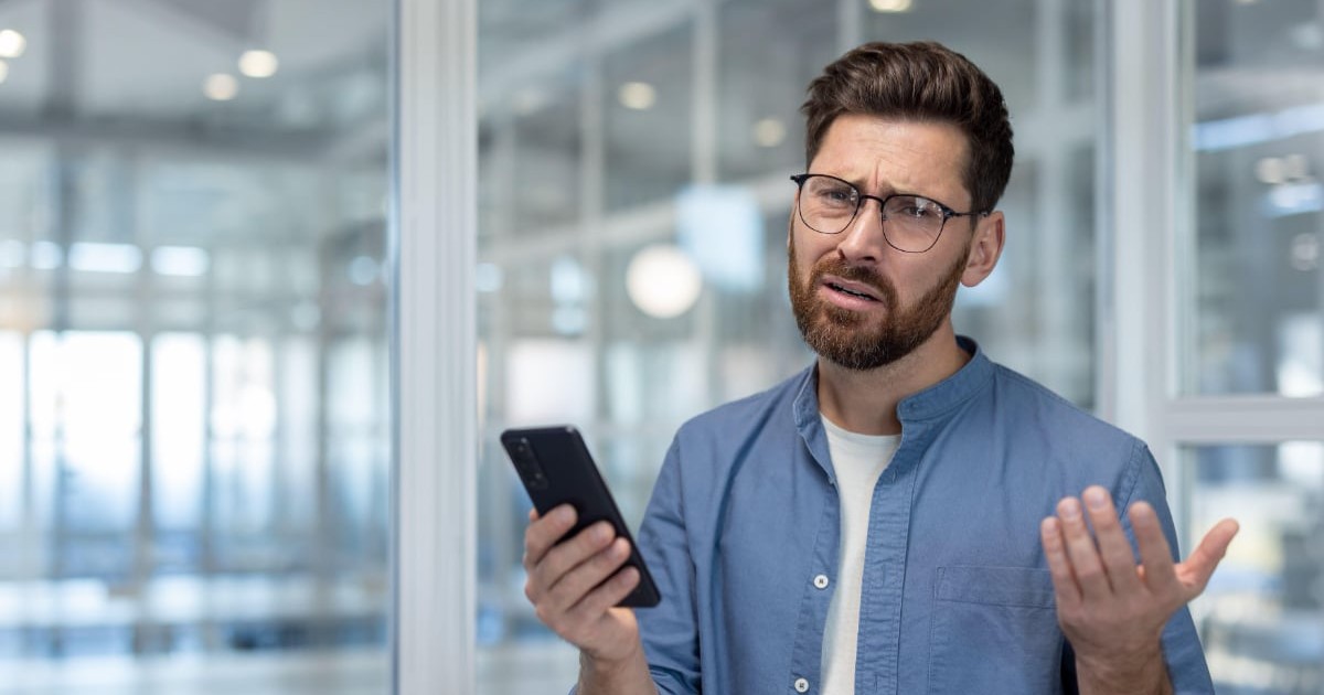 Un bărbat cu tricou alb, cămașă albastră, care ține un telefon în mână și este confuz