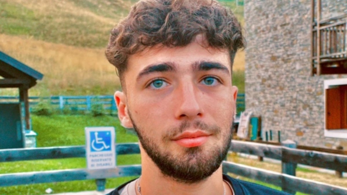 Imaginea prezintă un portret de aproape al unui tânăr cu părul ondulat, ochi deschiși la culoare și barbă scurtă, având în fundal o clădire din piatră și o zonă montană.