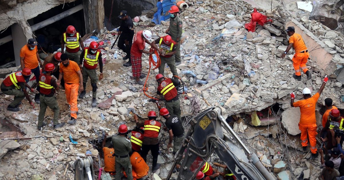 dărâmături – bucăți mari de beton, fier beton, praf și resturi de mobilier. printre ruine se află peste 15 persoane, majoritatea purtând căști de protecție roșii sau albe și veste reflectorizante. În partea de jos a imaginii, se vede brațul unui excavator.