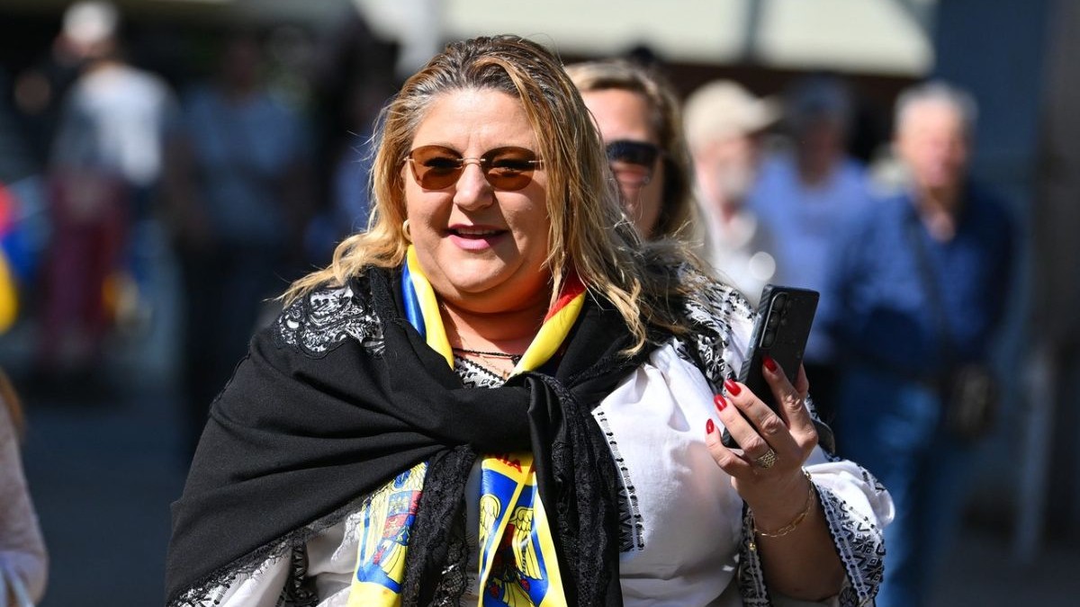 Diana Sosoacă intr-o ipostază pe stradă, rade, cu telefonul în mana stanga, care este ridicata, poartă o bluză alba cu model traditional romanesc si o eșarfă neagră legata la gat, oameni in spatele ei