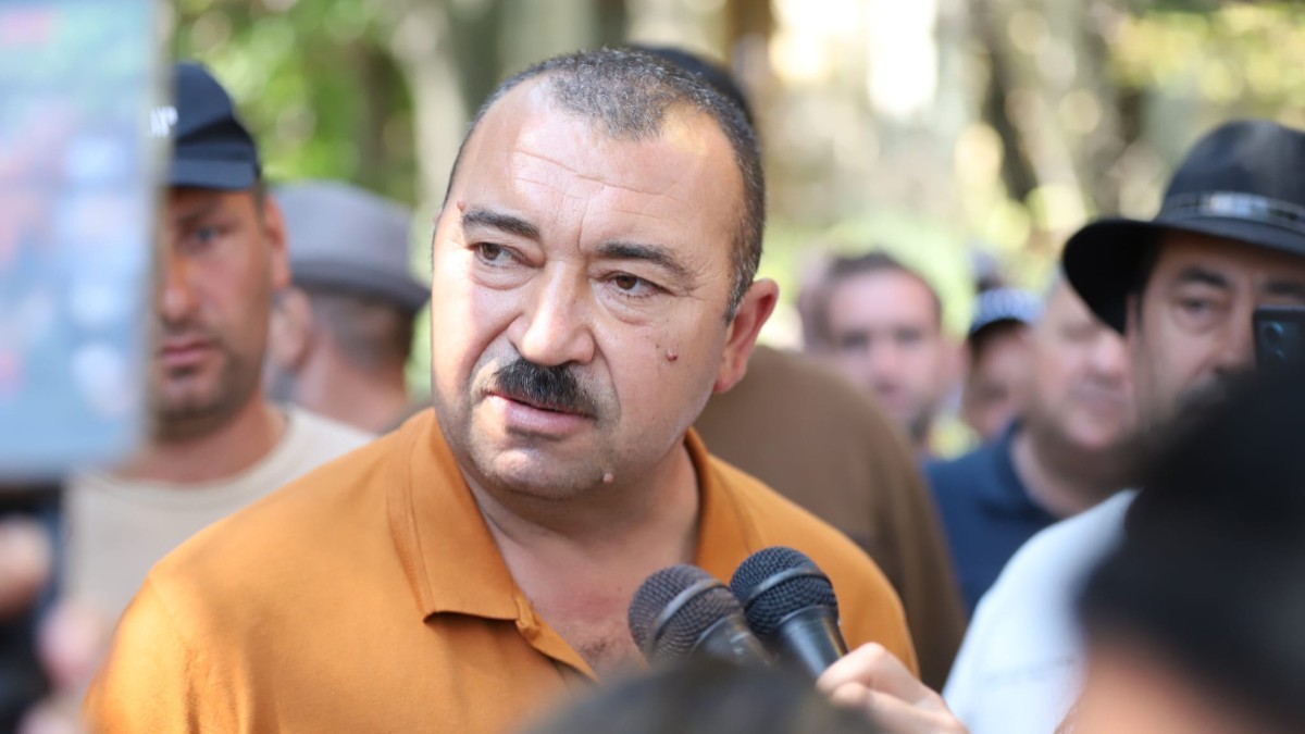 Un bărbat cu mustață vorbind în fața mai multor microfoane, înconjurat de oameni și jurnaliști