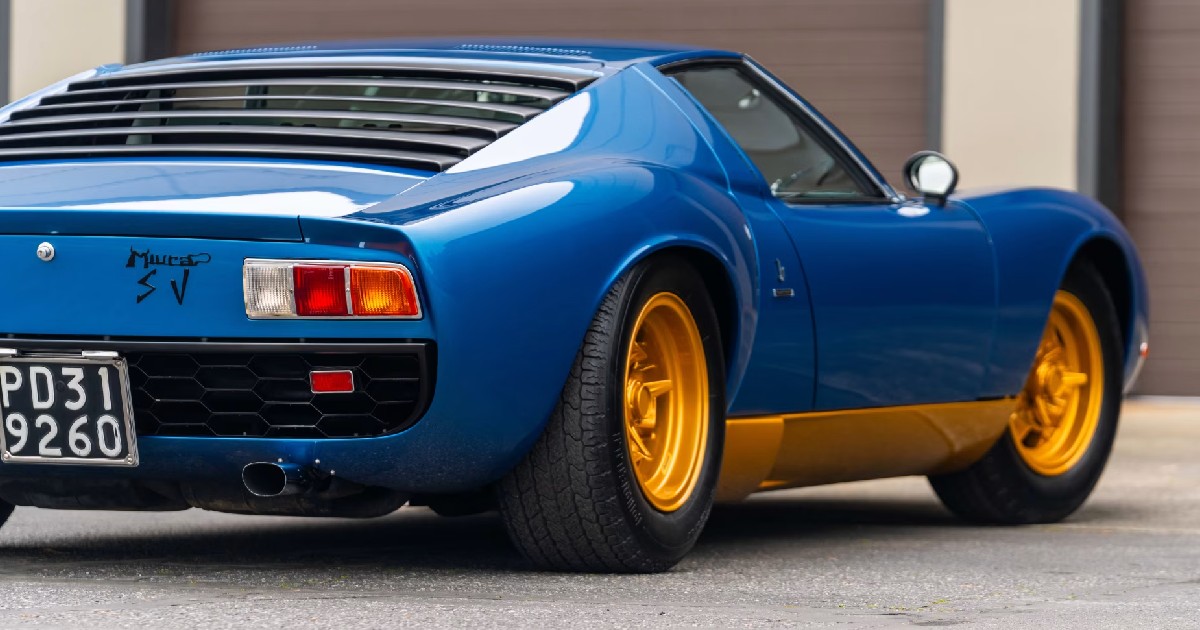 Imaginea prezintă partea din spate a unui automobil clasic Lamborghini Miura SV de culoare albastră, echipat cu jante aurii, parcat în fața unei clădiri cu uși de garaj.