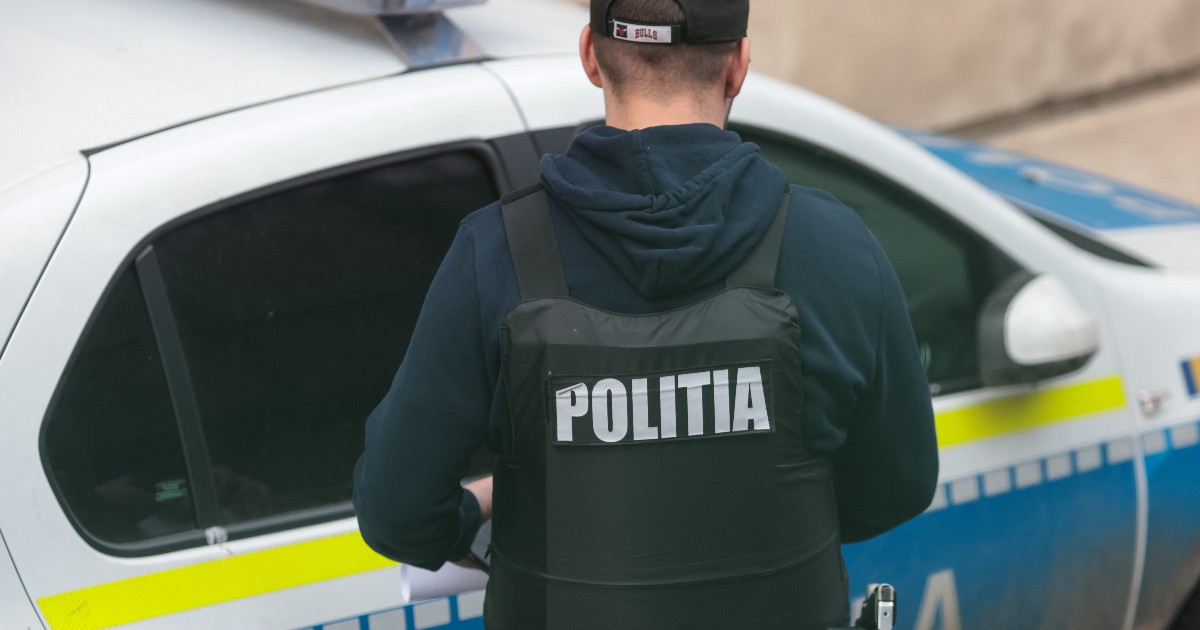 În imagine este prezentat din spate un bărbat ce poartă o vestă antiglonț inscripționată cu textul „POLIȚIA” și o șapcă neagră, stând lângă o autospecială de poliție alb-albastră.