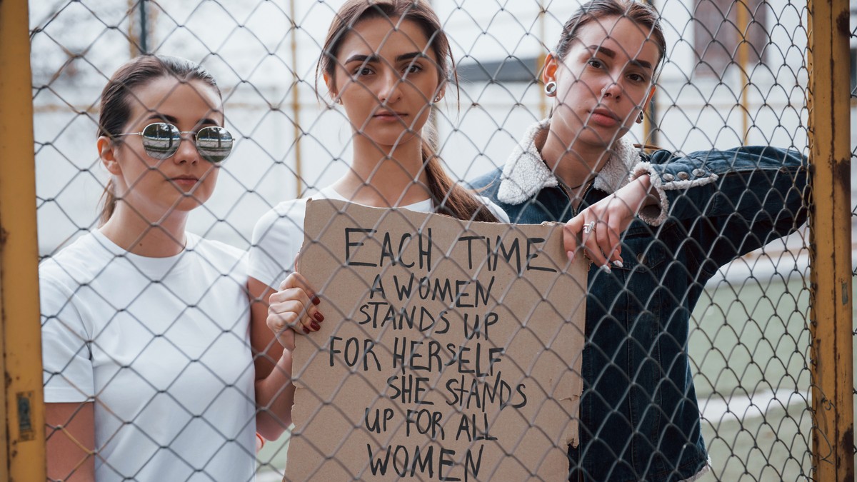 grup de femei care au un mesaj feminist scris pe un carton, in spatele unui gard