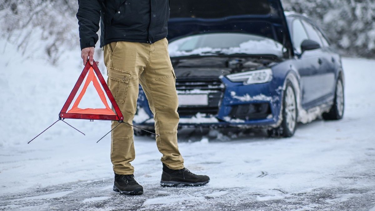 Fotografie cu un bărbat îmbrăcat într-o jachetă neagră și pantaloni de culoare bej, ținând în mână un triunghi reflectorizant, în fața unei mașini albastre care are capota ridicată, pe un drum acoperit cu zăpadă.