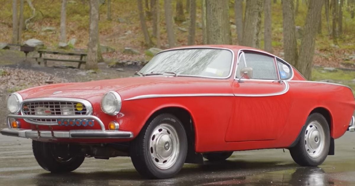 O imagine profil-față prezintă un Volvo 1800S clasic, de un roșu aprins, cu linii elegante și detalii cromate distinctive, parcat pe un carosabil umed care îi reflectă silueta, având în fundal o pădure rară cu o masă de picnic din lemn.