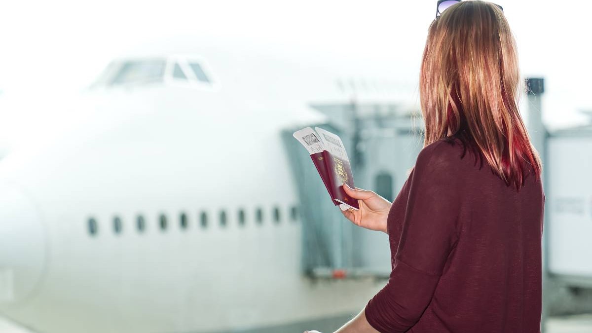 Femeie la geamul aeroportului , tine in mana pasaportul si biletele de avion, in findal se vede o aeronava pe pista